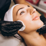 Woman on a treatment bed preparing for a 3D skin tech facial. Therapist wearing gloves wiping her face with cotton pads.