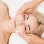 Young woman lying down with her eyes close receiving a 3D skin tech facial. Therapist wiping her face down with cotton pads.