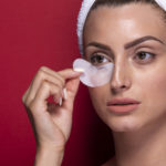 Closeup of a young woman wearing a towel applying an eye sheet mask to her skin.