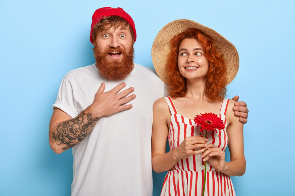 Excited happy man touches chest, impressed by good news, embraces girlfriend who holds red gerbera, have walk together, meet friends, isolated on blue wall. People, relationship, love concept