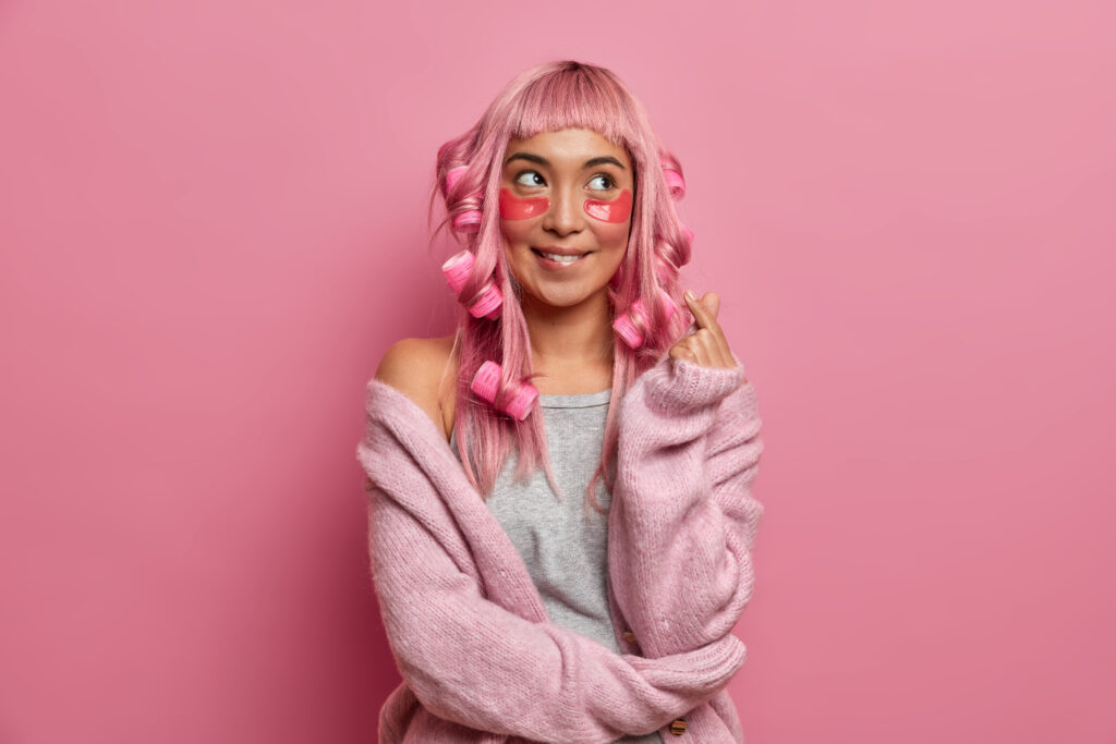 Photo of dreamy cheerful woman makes mini heart gesture korean symbol wears collagen patches under eyes, hair curlers, dressed in sweater, poses against rosy background. Wellness, hairdressing, beauty