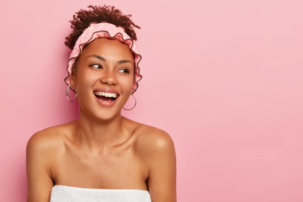 Hygiene, beauty and healthy lifestyle concept. Pretty smiling African American woman looks on right side stands wrapped in towel with naked shoulders wears shower hat enjoys taking bath and relaxation and treatments